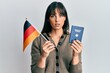 © Krakenimages.com - Young hispanic woman holding germany flag and passport relaxed with serious expression on face. simple and natural looking at the camera.