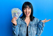 Young Hispanic Woman Holding 100 Romanian Leu Banknotes Celebrating Achievement With Happy Smile And Winner Expression With Raised Hand