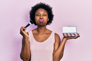 Poster - Young african american woman holding makeup brush and blush puffing cheeks with funny face. mouth inflated with air, catching air.