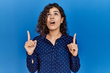 Poster - Young brunette woman with curly hair wearing casual clothes over blue background amazed and surprised looking up and pointing with fingers and raised arms.