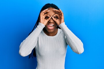 Poster - African american woman with braided hair wearing casual white sweater doing ok gesture like binoculars sticking tongue out, eyes looking through fingers. crazy expression.