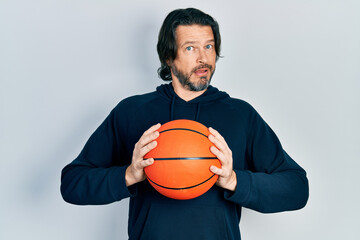 Canvas Print - Middle age caucasian man holding basketball ball in shock face, looking skeptical and sarcastic, surprised with open mouth