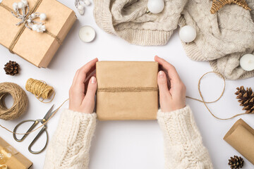 Wall Mural - First person top view photo of female hands in sweater wrapping craft paper giftbox with twine knitted scarf christmas tree balls and tools for handicraft on isolated white background