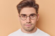Serious looking macho man with hairstyle and stubble wears round spectacles listens carefully to someone talking focused at camera isolated over beige background. Portrait of handsome guy indoor