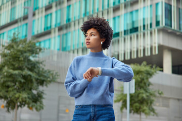 Wall Mural - Outdoor shot of thoughtful young woman with curly hair checks time on wristwatch waits for someone at street concentrated into distance wears casual jumper and jeans stands in urban setting.