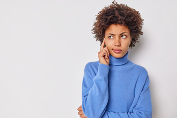 Photo of serious young curly woman keeps finger on temple tries to concentrate considers something contemplates about important thing wears casual blue jumper isolated over white background.