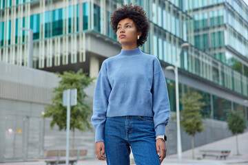 Wall Mural - Outdoor shot of thoughtful curly haired woman strolls in modern city concentrated into distance wears casual blue jumper and jeans enjoys free time at street poses against blurred background