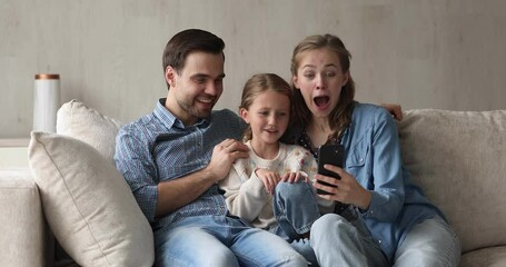 Wall Mural - Happy friendly family of three mom dad and small daughter cuddle on soft couch at living room watch amusing video on modern cell. Bonding parents and little girl enjoy funny clips online using phone