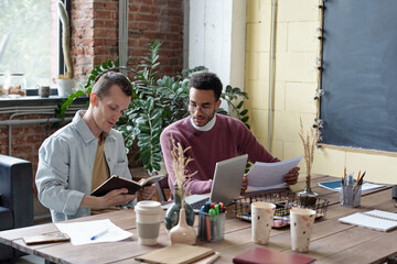 Poster - Two young intercultural businessmen in casualwear planning work while one of them looking at his notes in notebook