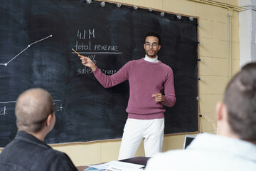 Poster - Young contemporary economist or coach explaining financial graph on blackboard while standing in front of audience