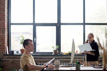 Poster - Young businessman computing by workplace while his female colleague with laptop speaking on mobile phone against window