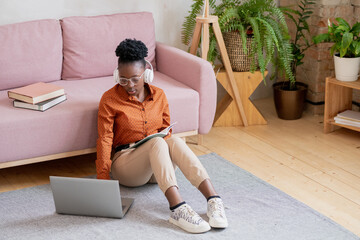 Aufkleber - Young serious black woman in casualwear and headphones sitting on the floor by couch in front of laptop during online lesson