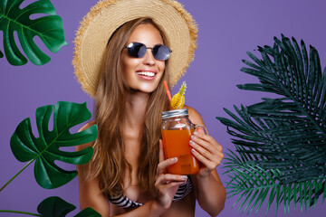 Wall Mural - Smiling girl holds fruit cocktail in sunglasses and straw hat while stands near tropical plants