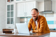 © Beton Studio - Young man working using laptop in kitchen in home interior. Smiling Freelancer works remotely from home. Happy man owner is doing paperwork using laptop indoors. Bald Caucasian 30s