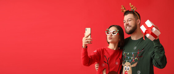 Wall Mural - Happy young couple in stylish Christmas clothes and with gift taking selfie on color background with space for text