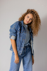 Wall Mural - young blue-eyed blonde woman with curly hair in a denim jacket and jeans stands on a gray background. concept of fashion and beauty in the studio. beautiful clean healthy skin. space for text