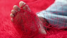 A Red Fishing Net Entangled In The Right Foot Of A Man Who Is Continuously Trying To Set Himself Free. Fisherman Using A Fishing Net Needle For Mending A Net.