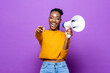 Smiling African American woman holding megaphone and pointing finger in isolated purple studio background