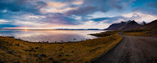 Panoramic Sunset Photo Of Road Leading Along Coast Of Lake To Volcanic Mountains. High Rocky Peaks Covered With Snow Layer Mirroring On Water Surface. Driver's Point Of View On Ring Road, Iceland.