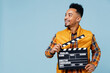 © ViDi Studio - Jubilant exultant young black man 20s years old wear yellow waistcoat shirt looking aside holding classic black film making clapperboard isolated on plain pastel light blue background studio portrait
