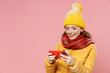 Gambling young woman 20s wear yellow jacket hat mittens use play race app on mobile cell phone hold gadget smartphone for pc video games isolated on plain pastel light pink background studio portrait