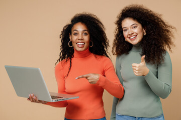 Wall Mural - Two vivid happy young curly black women friends 20s wear casual shirts clothes hold use work on laptop pc computer show thumb up like gesture isolated on plain pastel beige background studio portrait.