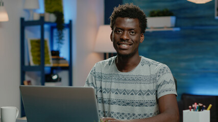 Poster - Close up of african american man working on laptop. Black employee doing remote work on internet while looking at camera and smiling. Business person of african ethnicity using device