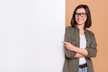 Wall Mural - Photo of cheerful lady direct forefinger placard board empty space wear specs khaki shirt isolated beige color background