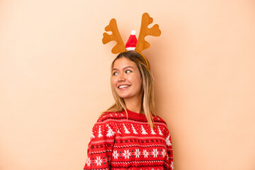 Wall Mural - Young caucasian woman wearing a christmas reindeer hat isolated on beige background looks aside smiling, cheerful and pleasant.