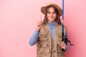 Wall Mural - Young caucasian fisherwoman holding a rod isolated on pink background showing number one with finger.