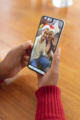 Wall Mural - Hands of woman making christmas smartphone video call with waving biracial men in santa hats