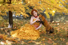 A Gentle Red-haired Girl Sits In An Autumn Forest In A Dress Made Of Leaves With A Red Fox