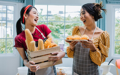 Two beautiful Asian women smiling with happiness and confidence, holding and presenting wooden container of basket of bread, croissant, baguette for bakery in indoor kitchen at home. Hobby Concept.