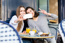 Young Woman Taking Selfie With Friend Showing Peace Sign At Cafe