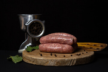 Canvas Print - Raw meat sausages on a wooden board on a black background. Homemade sausages in a natural casing next to a steel meat grinder. Meat products. Home cooking