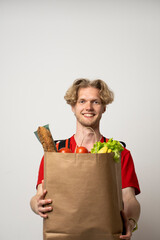 Sticker - Portrait of pleased delivery man in red uniform smiling while carrying paper bag with food products isolated over white background.