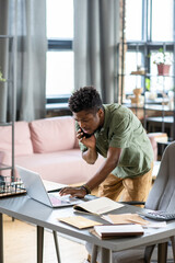 Poster - Serious young Blackman standing at table and checking information on laptop while talking by phone in office