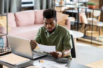 Concentrated young African American taxpayer in green shirt sitting at table with laptop and calculator and checking tax form