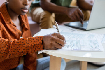 Poster - Close-up of Blackwoman in orange shirt filling tax papers at coffee table while her boyfriend using laptop