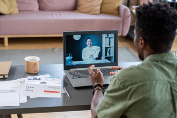 Poster - Over shoulder view of African American man sitting at table in living room and talking to financial consultant online