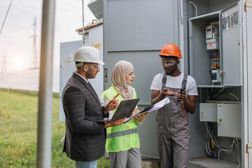 Wall Mural - Indian man and muslim woman with clipboard and laptop talking with african american technician at solar station. Multiracial people examining process of green energy production.