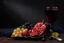 Still Life With Pomegranate, Black And Green Grapes, Middle Aged Styled Goblet With Vine, Knife, Napkin And Ceramic Or Porcelain Dish On A Dark Stone Table And Dark Background With Light Beam.