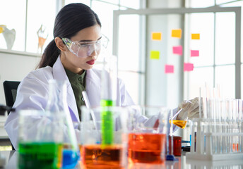 Wall Mural - Asian woman as chemistry expert pouring experiment liquid from beaker into glass tube with safety protection of goggles and glove at scientific lab