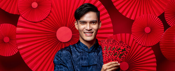 Wall Mural - Handsome smiling Asian man in traditional costume holding red envelopes in oriental style folded fan banner background, foreign text translation as great luck great profit