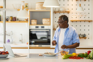 Senior African man standing in the kitchen, watching recipe on laptop and preparing a healthy meal.