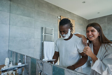 Wall Mural - Smiling lady examining foam on male face standing before mirror