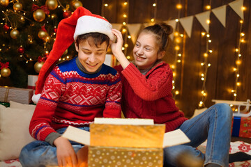 Portrait of a boy and girl in New Year decoration. They open a box, laugh and have fun. Holiday lights, gifts and a Christmas tree decorated with toys.