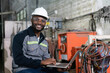 African American male engineer using computer laptop control robot arm welding machine in an industrial factory.