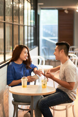 Poster - Smiling young Asian couple in casual outfits sitting at small table with drinks and chatting together in cafe