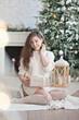 © Dina - Beautiful young woman with a gift box near christmas tree in the living room. Woman at home decorated for winter holiday season.